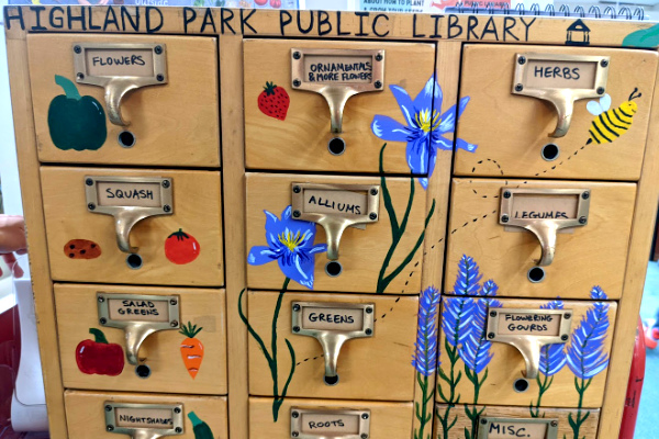 A wooden seed storage cabinet with small drawers, each labeled with a type of plant, is decorated with colorful paintings of plants, fruits, and vegetables.